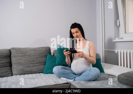 Una donna in attesa guarda lo schermo del telefono e indossa gli auricolari mentre siede sul divano nel soggiorno accanto alla finestra aperta. Foto Stock