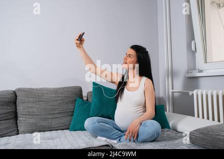 Una donna incinta che ama rilassarsi e divertirsi prendendo selfie. La mamma in attesa ama il tempo libero nel soggiorno. Foto Stock