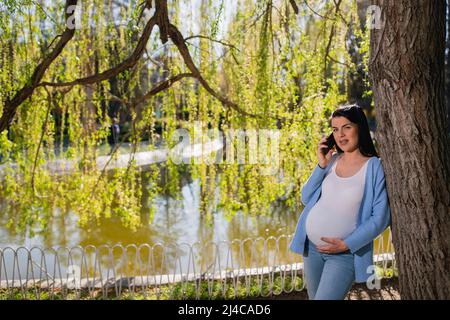 Una donna incinta sorridente con trucco parla sul suo telefono cellulare e guarda a destra la macchina fotografica mentre tiene il suo grande stomaco incinta con la sua mano sinistra. Foto Stock