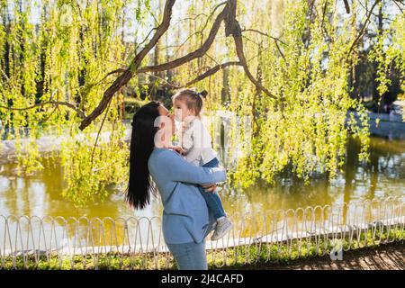 Una donna caucasica casual vestita tiene la figlia sollevata tra le braccia. Una bambina carina con ponytail bacia il naso della madre. Goditi la giornata di primavera Foto Stock