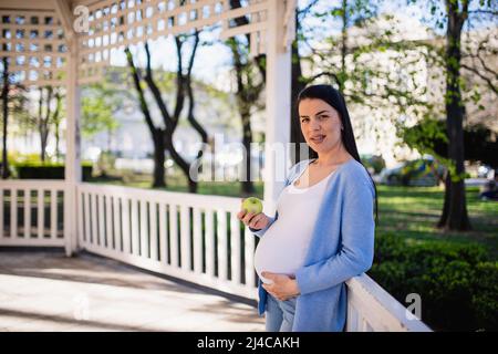 Una donna caucasica incinta dai capelli neri si appoggia contro una recinzione bianca nel padiglione di un parco cittadino e tiene una mela verde nella sua mano destra. Foto Stock