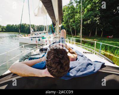 uomo adulto che si stende su un ponte a vela e prendere il sole durante le sue vacanze in una giornata estiva soleggiata. Yacht ormeggiato su un molo di legno su un lago Foto Stock