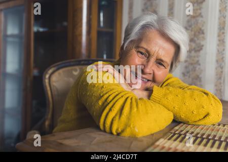 Donna caucasica anziana, pensionato gioioso, guarda la macchina fotografica e sorride mentre riposa le braccia su un tavolo di legno. Foto di alta qualità Foto Stock