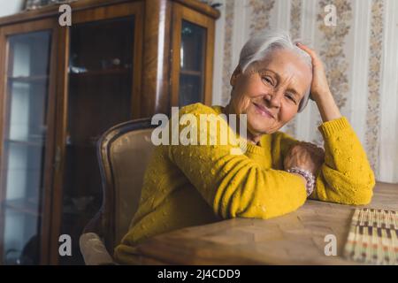 Felice della sua lunga vita signora grigio-capelli guardando la macchina fotografica e sorridendo mentre si rilassa da tavola di legno e toccando i suoi capelli. Foto di alta qualità Foto Stock