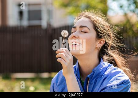 Primavera e felicità. Ritratto di una giovane donna che tiene un dente di leone e la tiene in faccia con un sorriso e occhi chiusi. Foto Stock