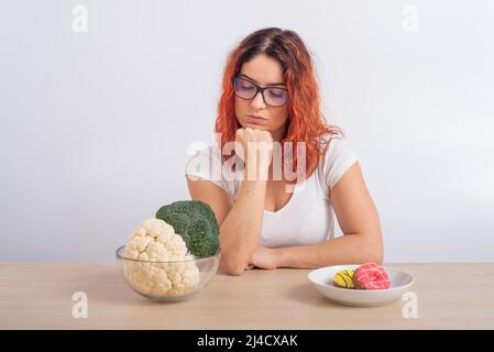 La donna caucasica preferisce cibo sano. Ragazza rossa sceglie tra broccoli e ciambelle su sfondo bianco. Foto Stock