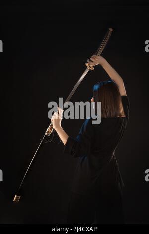 Una giovane bella donna in un kimono nero disegna un katana giapponese dalla sua scabbard, vista posteriore. Foto Stock