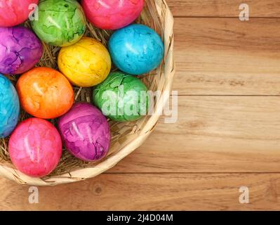 Vista dall'alto delle uova di pasqua colorate in cestino di paglia su sfondo di legno con spazio copia, Foto Stock
