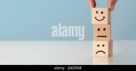 Valutazione del servizio clienti e analisi della soddisfazione concetti. La mano del cliente ha scelto felice faccia sorriso simbolo su blocchi di legno, sfondo blu. cop Foto Stock