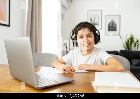 Concetto di formazione online. Scolaro concentrato che fa i compiti e ascolta la musica nelle cuffie, studiando a distanza da casa, utilizzando un computer portatile alla moda, preteen ragazzo scrivere attività nel notebook Foto Stock