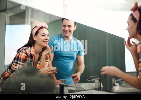 Allegro uomo brunette guardando bella ragazza applicare crema viso in bagno. Prepararsi insieme vicino allo specchio in bagno, Donna applicazione Foto Stock