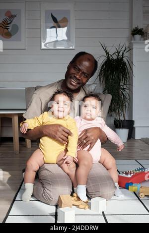 Ritratto di uomo afroamericano maturo allegro seduto sul pavimento in soggiorno a casa abbracciando i bambini gemellati sorridendo alla macchina fotografica Foto Stock