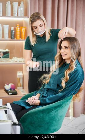 Giovane donna con capelli lunghi seduta in poltrona mentre parrucchiere fare acconciatura. Hairstylist femminile in maschera protettiva viso pettinatura capelli donna in salone di bellezza. Sfocatura del movimento. Foto Stock