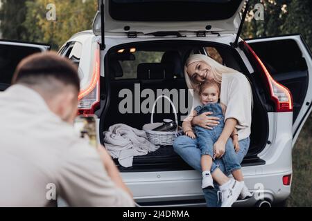 Marito fotografando sua moglie e figlia del toddler che seduta all'interno ha aperto il tronco di un'automobile di SUV, vacanza di viaggio della famiglia su strada Foto Stock