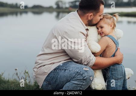 Il giovane padre abbraccia sua figlia e le diede il suo giocattolo morbido grande all'aperto vicino al lago Foto Stock