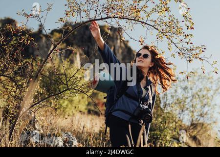 Ragazza giovane allegra di viaggio in occhiali da sole con zaino con tappetino da campeggio e fotocamera digitale su una cinghia che fa la sua strada attraverso il ispessito durante lei Foto Stock