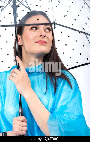 Scatto da studio di bella, felice e sorridente donna con lunghi capelli scuri indossando impermeabile blu e nascondendosi sotto trasparente con punti neri stile retrò Foto Stock