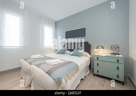 Camera matrimoniale con cassetti verdi pallidi, cuscini coordinati, testiera rivestita in tessuto grigio, carta decorativa sulle pareti e due finestre con tr Foto Stock