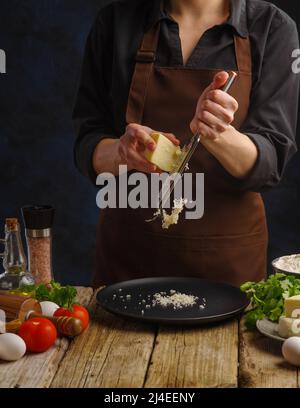 Lo chef prepara piatti a base di formaggio. Grattugiare il formaggio su una grattugia manuale. Ingredienti su un tavolo da cucina in legno. Ristorante, hotel, caffetteria, cucina casalinga, piccolo Foto Stock