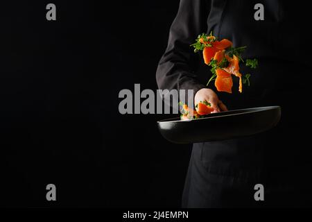 Lo chef prepara pezzi di pesce rosso in una padella - salmone, trota, tonno con verdure ed erbe su sfondo nero. Cibo congelato a bordo. Il Foto Stock