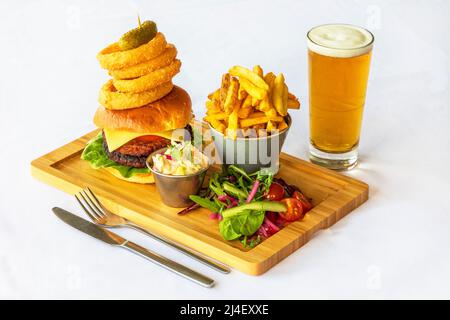 Server di legno con un rotolo e hamburger di manzo, anelli di cipolla, patate fritte coleslaw e insalata laterale e un bicchiere di lager accanto Foto Stock