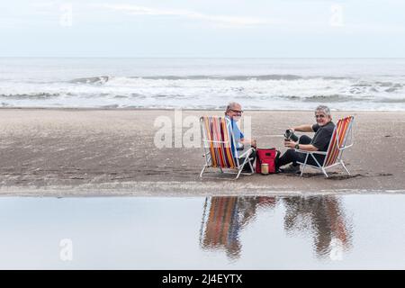 Due uomini di capelli grigi maturi seduti su sedie da spiaggia versando acqua in un compagno, sorridendo e guardando la macchina fotografica. Foto Stock