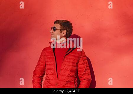 Giovane uomo in maglione rosso e giacca in piedi vicino al muro rosso Foto Stock