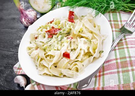 Pasta fettuccina con zucchine e peperoncino rosso caldo in salsa cremosa in piatto bianco su un asciugamano, aglio e una forchetta su fondo di legno Foto Stock