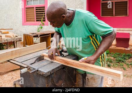 Un costruttore di cabinate con un pennel tra le sue dita si piega sul tavolo di una vecchia sega elettrica come sostiene un pezzo di legno 2 x 2 che usa sega t Foto Stock