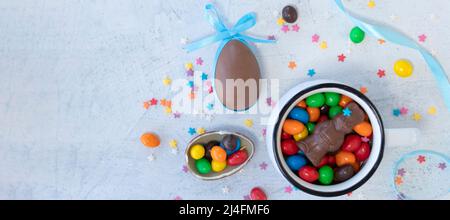 striscione con uovo di cioccolato con orecchie a nastro blu e varie caramelle di cioccolato in una tazza bianca su sfondo bianco. vista dall'alto, piatto. fuoco morbido Foto Stock