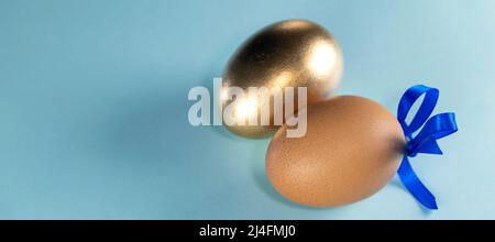 Striscione con uovo marrone con nastro blu e uovo dorato. Felice Pasqua concetto. Due uova con nastro su sfondo blu pastello con spazio di copia. Foto Stock