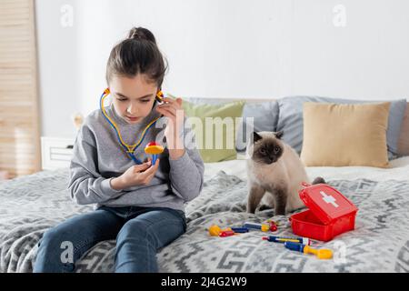 ragazza con stetoscopio giocattolo che gioca medico sul letto vicino al gatto e kit di pronto soccorso Foto Stock