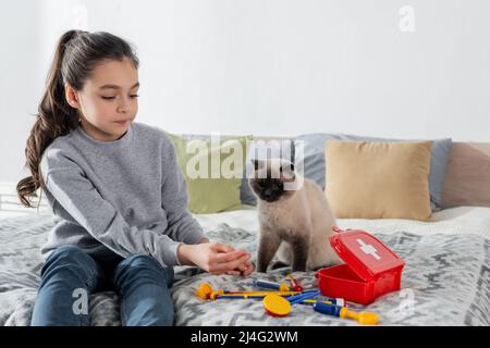 ragazza seduta a letto e medico di gioco con giocattolo set medico vicino al gatto Foto Stock