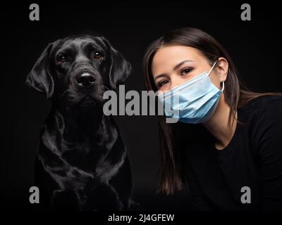 Ritratto di un cane Labrador Retriever in una maschera medica protettiva con una donna proprietario. La foto è stata scattata in uno studio fotografico Foto Stock