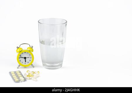 Un bicchiere d'acqua, una sveglia gialla e pillole su sfondo bianco Foto Stock