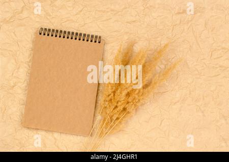 Blocco appunti vuoto con carta kraft su sfondo beige crolverato con erba di pampas. Modulo per congratulazioni o inviti. Foto Stock