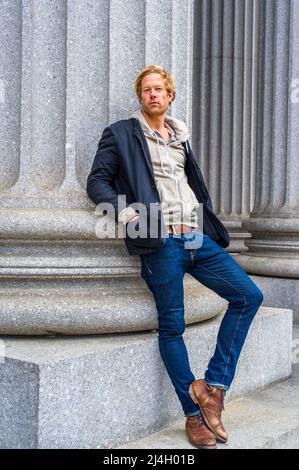 Vestendo in una giacca nera all'esterno e una giacca con cappuccio all'interno, jeans e stivali, un giovane ragazzo bello è appoggiato contro una colonna all'esterno di un ufficio costruire Foto Stock