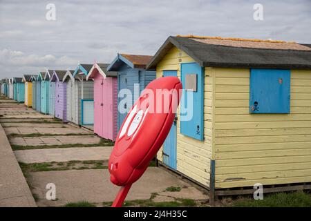 Una fila di capanne di spiaggia dai colori vivaci sul lungomare / passeggiata a Brightlingsea, Essex. Anello di salvataggio rosso in primo piano. Foto Stock