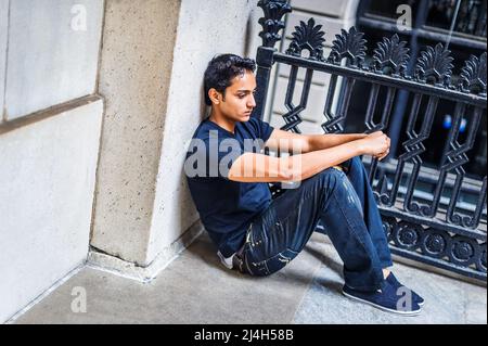 Solitario asiatico americano teenager profondamente pensando fuori a New York City, indossando maglietta nera, jeans alla moda rotto, scarpe di stoffa, seduta su grou Foto Stock