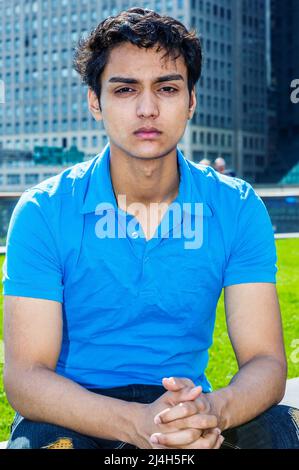 Ritratto di 18 anni adolescente indiano-americano a New York City. Giovane studentessa collegiale maschile che indossa una camicia a manica corta blu, seduta di colore verde Foto Stock