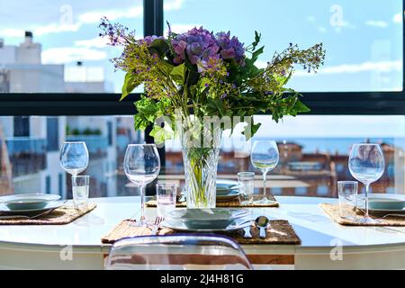 Bella disposizione tavolo con bouquet di fiori in vaso, posti serviti con stoviglie, mare e case tetti vista attraverso la finestra Foto Stock