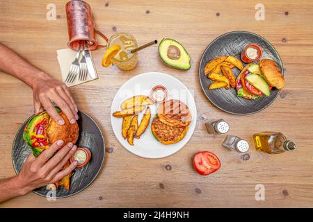 Mani che tengono un hamburger accanto ad altre tre con avocado, manzo, patatine fritte a spicchi, pomodoro e pepe, olio e sale Foto Stock