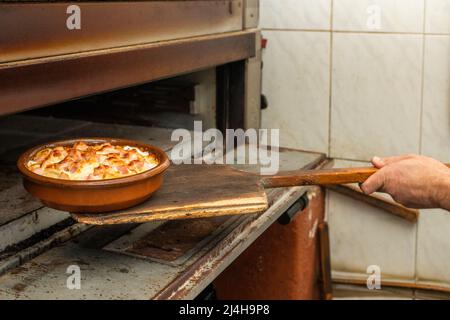 Utilizzando una pala di legno, togliendo alcune patate con pancetta e panna dal forno in una pentola di argilla Foto Stock