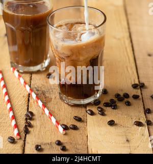 Caffè ghiacciato in grandi bicchieri trasparenti, versato sul latte, con chicchi di caffè su sfondo di legno, bevanda rinfrescante estiva Foto Stock