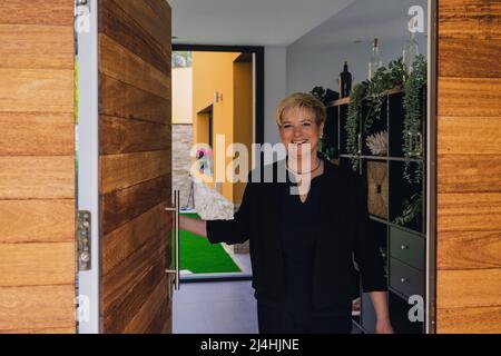 Donna matura sorridente con capelli biondi che aprono la porta ai visitatori. Donna della sua piccola impresa che apre la porta. Foto Stock