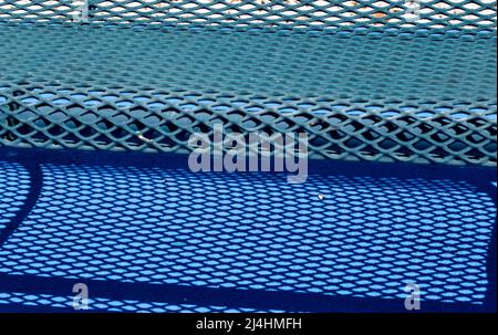 Un banco in acciaio a rete dipinto di blu proietta una forte ombra su un marciapiede dipinto di blu. Foto Stock