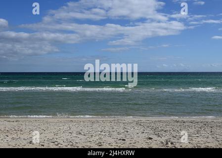 Spiaggia di sabbia con molte impronte, con onde basse e dolci dal mare che si infrangono verso di essa, durante una giornata di sole con nuvole chiare nel cielo Foto Stock