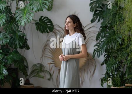 Giovane donna felice giardiniere in piedi contro lussureggianti monstera Deliciosa houseplants in giardino di casa Foto Stock
