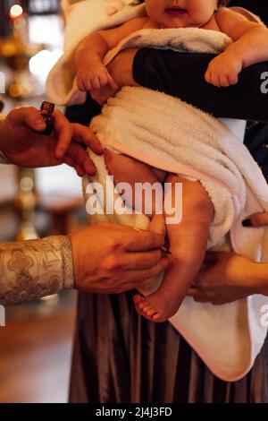 Foto ritagliata di sacerdote in cassone che tiene olio santo per unzione con piedi di spazzola del bambino durante la cerimonia del battesimo. Foto Stock