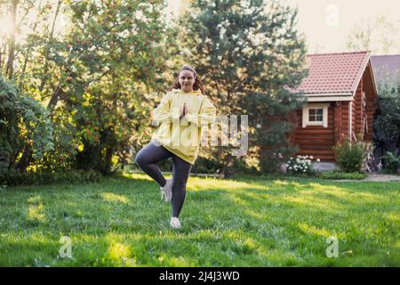 Donna che si bilancia su un piede di erba fresca facendo esercizio yoga indossare abiti sportivi guardando la macchina fotografica sul cortile con casa di campagna e alto Foto Stock